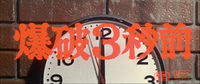 Blood red titles calligraphy in Japanese are superimposed on a close-up of a clock, set to 3 seconds before 12 o'clock, mounted on a red brick wall.