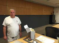 Man in white T-shirt standing beside a desk with an open box of archival documents.