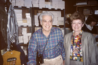 The two smile as they look around the office and shop. This color photo shows them in the mail order department room with shelves stacked with postcard inventory.