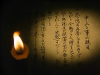 A page of parchment has black calligraphy written on it, with a candle casting a candlelight circle on it.