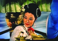 A color film still featuring a medium close-up of a woman sitting in a boat. She is gazing to her right and is holding a red scarf in her right hand.