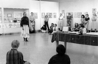 Black-and-white photo. Brooks performs in a long, embroidered skirt and shawl in the middle of the gallery. Guests watch, seated or standing along the walls. Displays in the background.