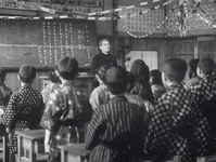 The foreground is filled with the backs of elementary school pupils looking at their instructor. The instructor faces them next to a chalkboard. Text is written on the board with chalk. There are calligraphic strips of paper near the ceiling in the background.