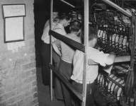 1930s era photograph of five men lined up behind a row of hand levers attached to electrical dimmers, “No Smoking” sign to the left side.