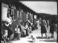 Refugees sitting outside makeshift homes, man playing guitar.