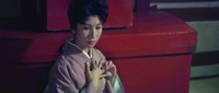 Close-­up on a seated Mrs. Iwata in her kimono, just Left of center, with eyes downcast and making a covert signal with her hands and the large jade ring. A large, wide, painted red chest, below a smaller chest, dominates the simple, shallow background.