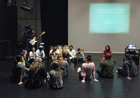 Approximately a dozen dance students and members of the group Quetzal sit in a circle on the floor of a rehearsal space. Various musical instruments fill out the scene.}
