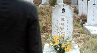 The back of a man in a black jacket obscures the left side of the frame. The right side of the frame reveals yellow flowers planted in front of a gravestone with red calligraphy written on it.