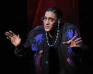 Jones in a colorful patterned sweater with shoulder pads, wearing oversized 1980s-­style glasses attached to a plastic beaded chain. Her hands are out to either side, as if trying to get someone to listen to her.