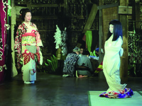 A busy, multi-­planar backstage scene. Though at only a slight angle, the camera captures the stiff child playing the snow queen (screen Left), in front of what appears to be a wooden stage door, as well as the backstage wings where Shinako (Left middle-­ground), in front of a curtain and various stage architecture, tries to question the girl. Between them, background, are some adults seated in a circle. Behind them and also to their Left and Right are old props, scenery pieces, curtains, etc.