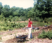In a T-shirt and long pants, Cohen stands, smiling, with a shovel and a wheelbarrow full of compost next to the double dug beds. Garden, meadow, and forest beyond.