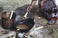 A color photograph of a figure cooking balsam fir pitch in an iron pan over an open flame.