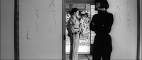 Oriko stands in the midground just inside the opening in the shoji with black calligraphy painted on it, in black and white cinematography. Behind her is the tall brush of a garden. In the foreground is a woman in all black to Oriko's right at the edge of the opening in the shoji, looking to the left with her arms crossed.