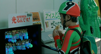 A boy in a shop is outfitted in a spandex jumpsuit and helmet with a black English numeral printed on them. Signs on the wall in front of him have red and green calligraphy printed on them.