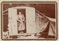 Carr stands at the door of her trailer, smiling. She wears a long dress of printed fabric and a vest. The trailer has a tent-like area on the right; a broom hangs to the left.