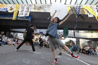 Dancers holding umbrellas perform in the street. The female dancer in the foreground stands on one foot and stretches her arms above her head. People sit or stand on either side to watch. Banners with Chinese text hang from the overpass behind the dancers