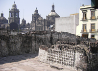 The Templo Mayor is made of gray, somewhat crumbled stone walls, one of which holds rows of carved skulls. The ornate cathedral stands in the background.
