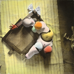 The hand-tinted black and white photograph from The Games of the Doll series (1938-1949) by Hans Bellmer shows the doll’s head, abdominal sphere, thigh, and two pelvises all broken apart and curled around the broken parts of a caned chair seat and leg. All these fragments are assembled on a tablecloth on the floor, adding to them a white hair bow. The bow accentuates the triangular layout of the composition, while the pink, red, orange, green and blue tints on the body parts give them the appearance of fruit.