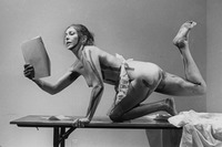 Carolee Schneemann kneels on a table with her right leg raised in the air while reading from Cezanne: She Was a Great Painter. She wears a white maid’s apron and has paint lines along her body.