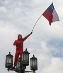 Protests in Santiago, Chile, in 2019. An individual stands at the top of a four-­lamp streetlight, wearing a red jumpsuit and red hoodie with the hood up. A mask covers their face. The mask is white with raised eyebrows and wide eyes indicative of surprise. Under a large nose, a long, thin, black moustache points up at the edges, with its points touching the bottom of the eyes. The mouth points slightly down at the sides. The individual’s right hand is pressed against their heart and their left arm holds up a large flag of Chile on a thin pole.