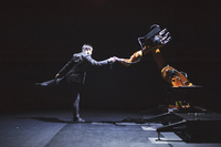 Figure 16.1. On stage, a man in a suit poses on one leg, the other lifted behind, grasping “hands” with an orange and black industrial robot. Their poses roughly mirror each other.