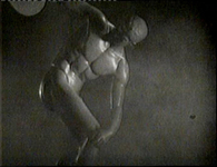 A statue of a naked olympic physique poses in darkness and fog, in black and white cinematography.