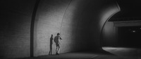 Long Shot of the Left wall (and partial unfinished ceiling) of an underpass tunnel. Hanada (center Right), a small figure in a white suit straggling by the tunnel wall. A strong light from the Right illuminates the center of the wall, casting Hanada’s strong shadow upon it. In front and behind, the lower parts of the wall in shadow. Outside the sphere of the tunnel exit (Right) the road climbs up and under a small traffic bridge.
