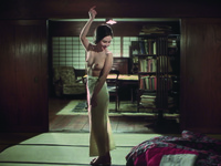 A deep focus composition, with Sono standing, middle-­ground and center, in front of the threshold between the sitting room (with a large settee protruding at an angle the bottom Right of frame) and what appears to be Nakasago’s Westernized study, with bookshelves, chairs and a large table. Topless, with a suddenly gay expression, Sono tosses her arm over head and snaps her fingers, as if making a dancing pose.