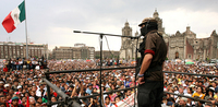 Marcos, wearing a full face mask, addresses Mexicans in a completely packed Zócalo. He is on the east side of the square on an elevated platform, speaking into a microphone.