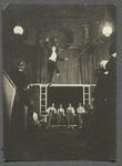 Photograph of Goluvtin crossing over the audience's heads on an inclined tightrope wearing a tuxedo, with bare feet, holding a parasol. Also visible is some of the detail on the walls and ceiling of this ornate room in the mansion that before the Revolution had belonged to the wealthy Morozov family.