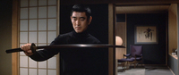 A man in a black turtleneck examines a sword. In the background room, calligraphy can be seen on the wall.