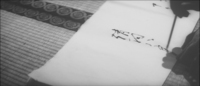 Closeup of a man writing out a name by brush, on paper that sits on tatami.