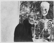 The Photograph of Hermine Moos with the skeleton of Oskar Kokoschka’s Alma Doll depicts the young doll maker clad in a black winter coat with a fur collar, posing next to a life-size skeleton made of papier mâché in an outdoor setting that looks like a balcony, most likely in her parents’ Munich apartment. She is holding the doll’s upper arm bone with her right hand, tilting her head towards its shoulder bone, while fully smiling at the camera.