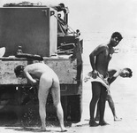 3 IDF soldiers shower in the desert. 1 pale nude soldier faces away from the camera. 2 dark Mizrahi soldiers smile and cover themselves.