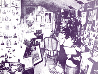 Willard, barely seen, sits in a wicker chair, her back to the viewer. The desk is piled high and her cluttered office is crammed with photos tacked on the wall and framed photos on the floor, unhung.