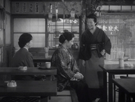 Women converse in a restaurant with black calligraphy on a variety of banners, in black and white cinematography. A shop sign in white calligraphy can be seen across the street.