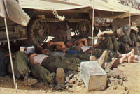 Color photo of 10 IDF soldiers in half uniform stretched out and napping next to one another under the shade of a tank in the desert.