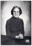Studio portrait of Gleason with a warm smile, wearing a crew neck sweater and large, square-rimmed glasses, hands folded in front of her on a table. WWLW logo in lower corner.