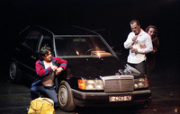 A woman in leopard print gropes man lighting a cigarette leaning against a car. A man in a tracksuit crouches on the other side of the car.