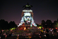 In stark relief to the dark sky behind it, a bright light illuminates a monument marked by graffiti in multiple colors, projecting onto it the face of a Black man, his features both obscuring and blending in with the stones of the monument. Atop the monument is a statue of a figure on horseback. Projected in white light on the horse are the letters “BLM.” Just below the horse and above the forehead of the man’s image is the phrase “Black Lives Matter.” Several people appear at the base of the monument.