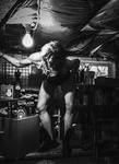 Black-and-white photograph of a female figure positioned on top of a workbench in a crowded basement workshop space.