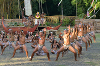 Photograph showing an ensemble of Tahitian dancers performing outdoors. A stage replica of a double-hulled voyaging canoe is in the background.