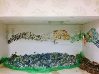 An installation. A colorful wave of plastic debris is on the walls. Plastic jellyfish and wood containers hang in the air. On the ground are a sea of plastic bottles and netting and cliffs made of cardboard.