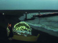A ghostly composition of the dimunitive Toyoko, Screen Left, in funeral dress standing on a twilight beach cove next to an oversized boat that has inside it an almost fantastically oversized funereal bouquet of white flowers. Off the shore, a line of innumerable dock posts stick out of the water with a votive candle on each post.