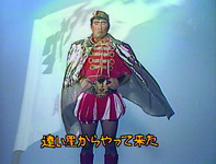 Color still of man (Kitano Takeshi) in red prince-­like costume, silver cape and make-­up. Silhouette of flag overlays man. At bottom of screen, text transcribes lyrics from song.