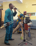 Two musicians perform in a carpeted room. One in the foreground sings into a microphone while holding a trumpet, wearing a traditional African outfit with a shell necklace and sunglasses. The other musician plays a long, tall wooden drum while singing. He wears a traditional African outfit with a cloth cap. Other drums and shakers are seen on the floor in the foreground.