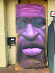 Image is a photo of a plywood mural in honor of African American man's face, likeness of George Floyd, plywood propped against yellow brick wall