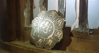 Image of a silver, flower-shaped plate with gold calligraphy engraved into it as a pattern. The plate rests against a window on a windowsill.