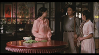 Image of three people standing around a round wooden table at a restaurant. One person is leaving and saying goodbye from the doorway. The other two people look at him. There is a bowl of greens on the table and, through a window in the deep background, red and pink banners with black calligraphy on them.