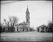 Black-and-white photograph of entrance to Cave Hill Cemetery in Louisville. Entrance includes gatehouse with tall clock tower. The road in front of the entrance appears to be brick or cobblestone, and there are trees on either side of the entrance.